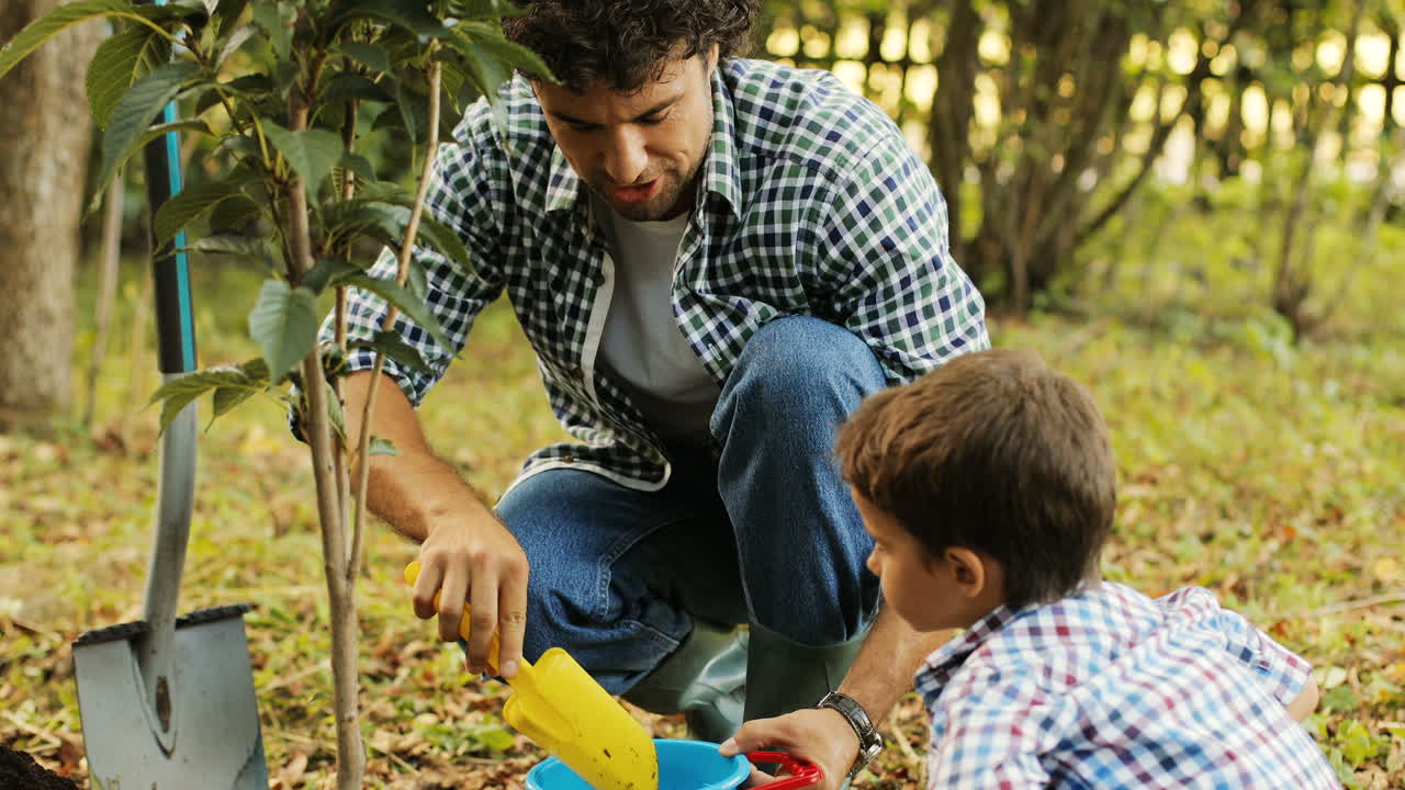 primer plano. retrato de un niño y su padre plantando un árbol. papá pone la tierra con la pala de juguete en el cubo de juguete. hablan y sonríen. fondo borroso