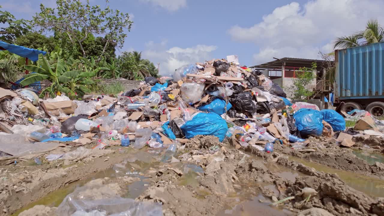 una toma de grúa de un vertedero de basura en un día soleado con agua sucia, plásticos desperdiciados, contenedores abandonados, árboles y nubes