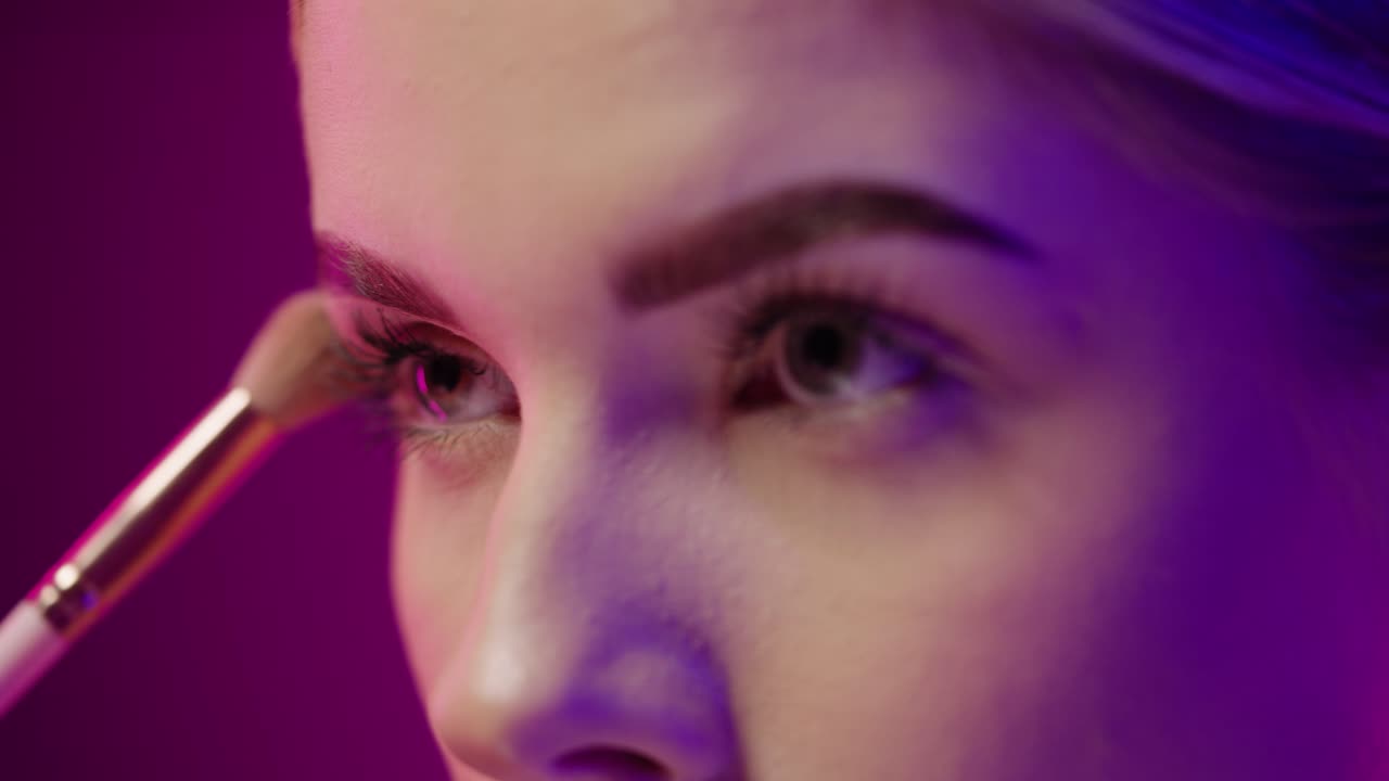 primer plano de una hermosa mujer joven poniendo maquillaje con un pincel de polvo y maquillaje en él mientras ella está en polvo su cara para la noche frente al fondo negro púrpura en cámara lenta
