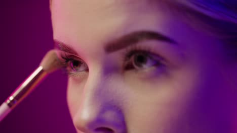 Close-up-of-a-beautiful-young-woman-putting-on-make-up-with-a-powder-brush-and-make-up-on-it-while-she-is-powdering-her-face-for-the-evening-in-front-of-purple-black-background-in-slow-motion