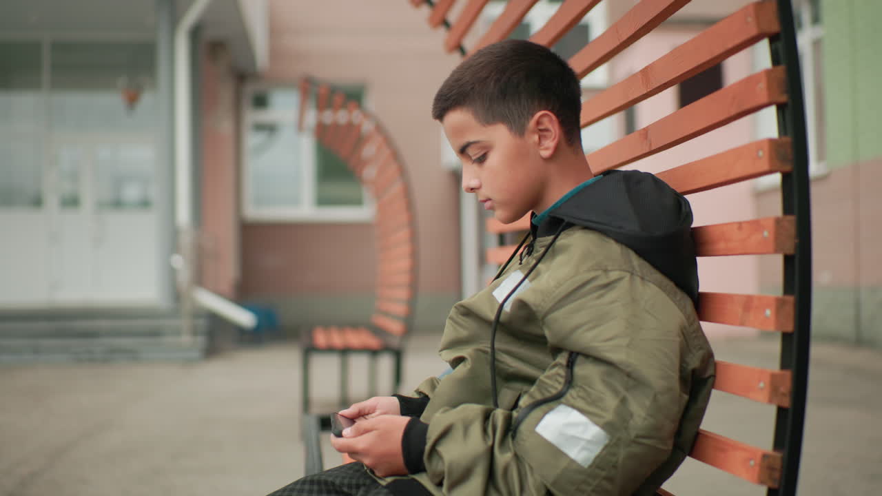 Vista laterale di un ragazzino con felpa e giacca seduto su una panchina, concentrato a giocare con il telefono vicino all'entrata della scuola con sfondo sfocato, creando un'atmosfera calma e rilassata
