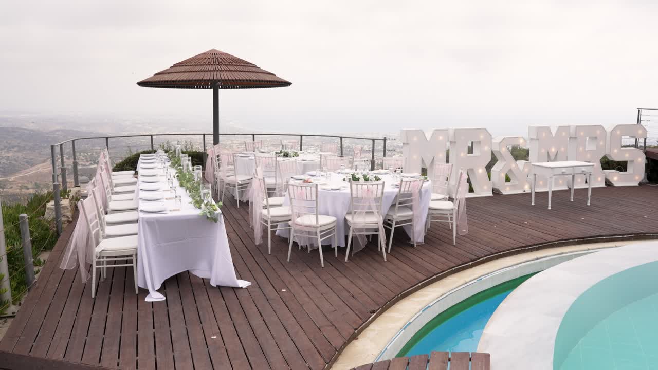 Tables are arranged tastefully on a terrace, set up for a wedding celebration. The area showcases a lovely pool and stunning scenery in the background
