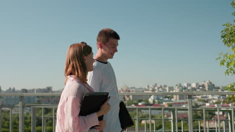 young adults walking near iron railing, man with tattooed arm conversing warmly with smiling woman holding notebook, background featuring greenery and expansive cityscape under clear blue sky