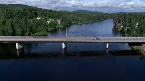 a car driving on a small bridge over a river surrounded by trees with mountains in the far background