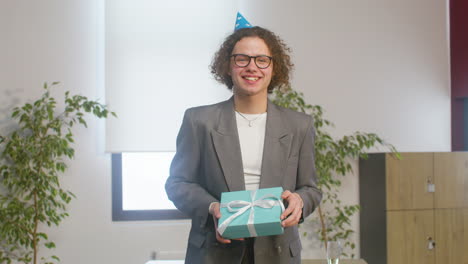 retrato de un niño rizado feliz con sombrero de fiesta sosteniendo una caja de regalo y mirando a la cámara durante una fiesta en la oficina