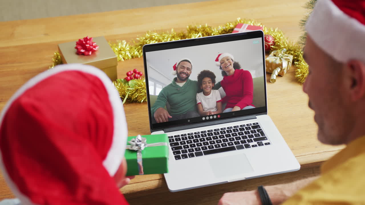padre y hijo caucásicos con sombreros de santa usando una computadora portátil para una videollamada de navidad con la familia en la pantalla