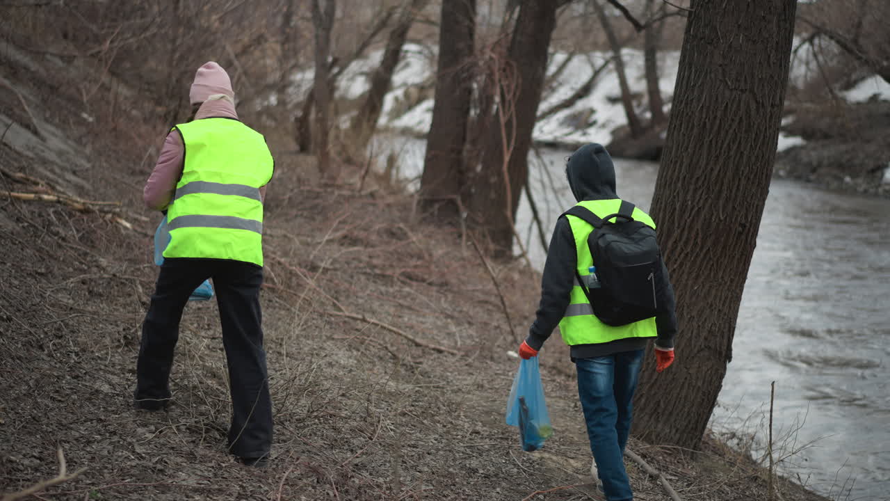 Two volunteers in reflective safety vests picking up litter along riverbank during environmental cleanup, carrying blue plastic bags while removing waste from natural area to protect wildlife