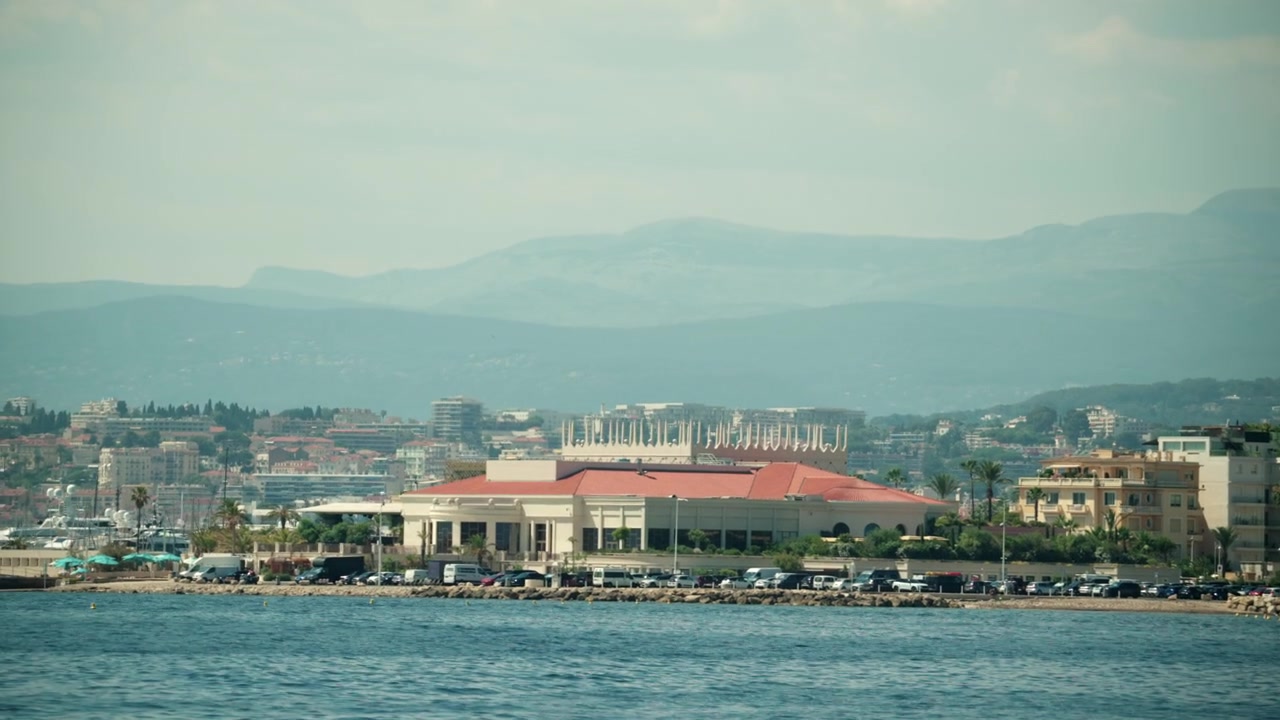 Paesaggio urbano di Cannes sulla Costa Azzurra, con edifici lungo la costa e montagne visibili sullo sfondo.