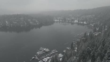 white snow falling down over green pine trees and marina with small boats docked in deep cove bay in british colombia on a cold cloudy winter day