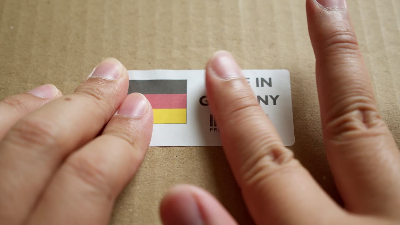 Mani che applicano l'etichetta con bandiera "MADE IN GERMANY" su una scatola di spedizione con codice a barre del prodotto di qualità premium.