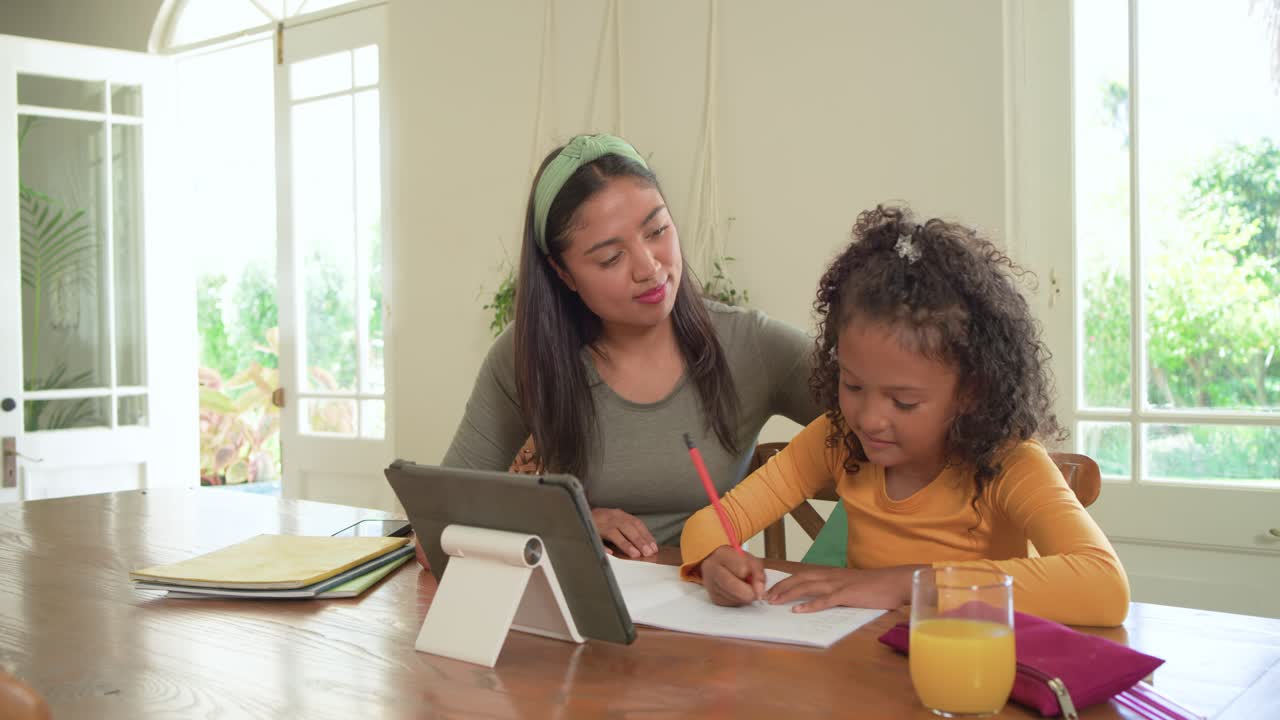 Madre guiando a su hija de la tableta al papel en la mesa de casa, señalando mientras la niña practica la escritura.