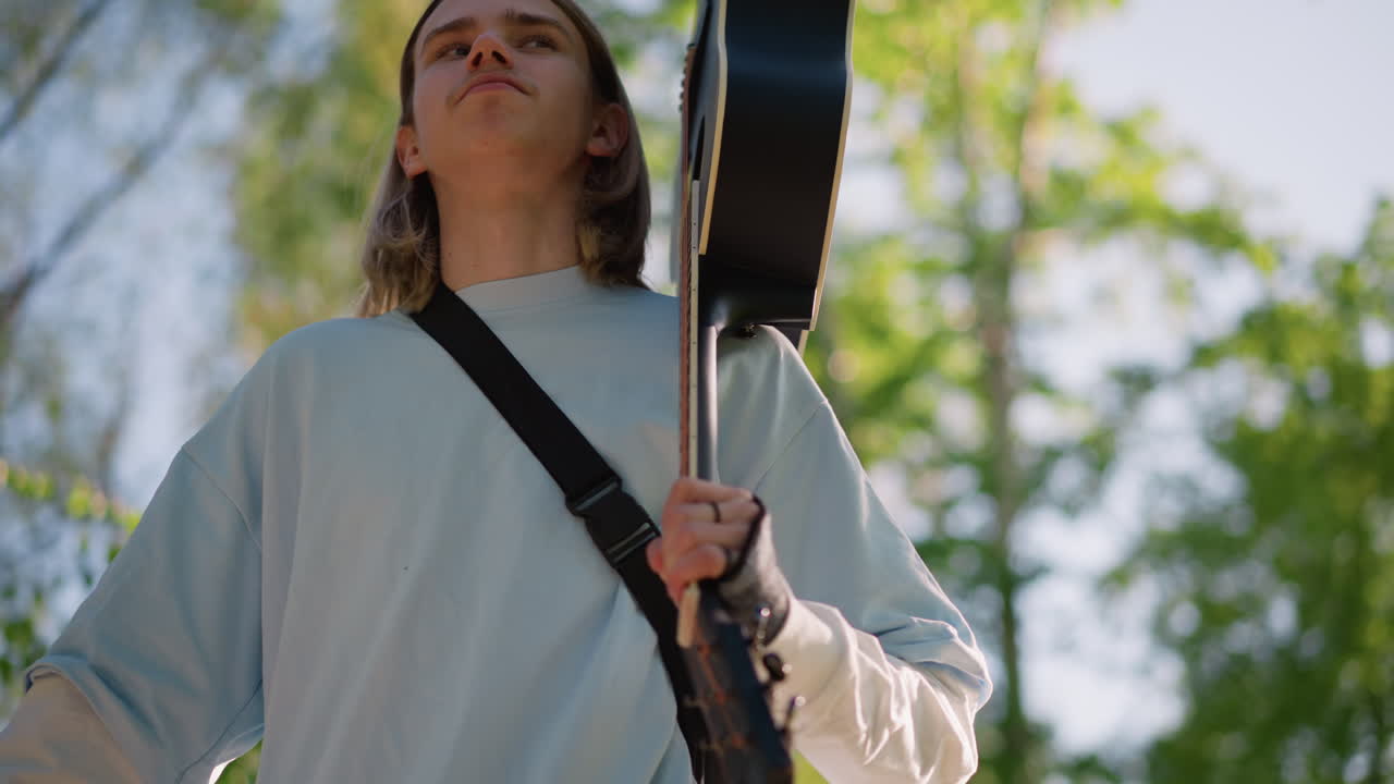 Joven guitarrista blanco con funda de guitarra atravesando un parque frondoso, con la cabeza inclinada hacia el cielo, energía de artista callejero seguro y aire de compositor de gira, fondo con luz solar moteada, fotos espontáneas tomadas a mano, alegre