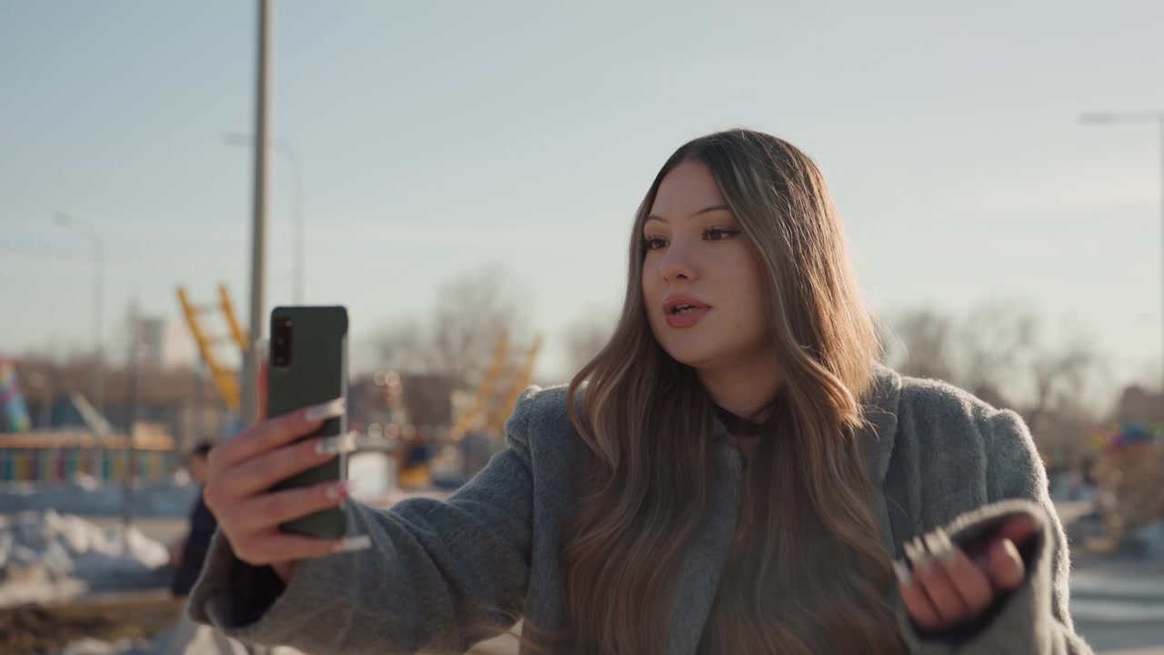 Pretty asian woman taking selfie outdoors, winter coat, golden hour sunlight, holding smartphone, posing with subtle smile, urban street backdrop, soft bokeh, confident expression, fashion aesthetic