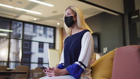 Video-of-thoughtful-caucasian-businesswoman-in-facemask-using-smartphone-in-lounge-area-at-office