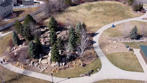 drone flying in circles around a public rock park with playground, revealing suburban middle class american neighborhood, while children play, on a sunny spring day after covid-19 lockdown lifts