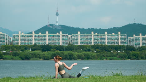 Fitte-Junge,-Flexible-Frau,-Die-Beine-Und-Körper-Streckt-Und-Übungen-Auf-Der-Matte-Im-Freienpark-Am-Fluss-Macht,-Mit-Der-Wunderschönen-Landschaft-Des-Namsan-N-Seoul-Turms-Im-Hintergrund