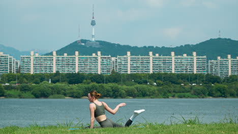 fit young flexible female stretching legs and body doing exercises on mat at outdoors park by the river with beautiful landscape of namsan n seoul tower in background