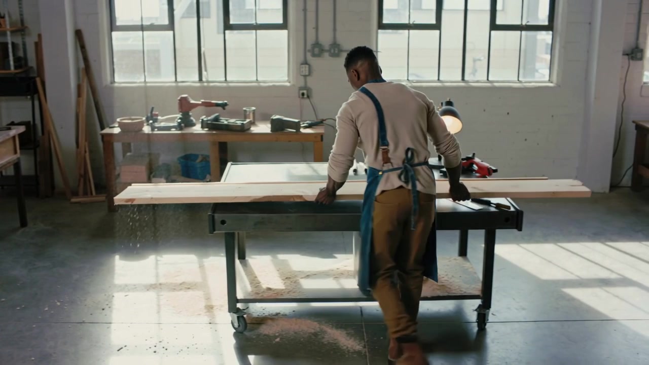 Een werknemer in schort gaat een houtwerkplaats binnen met twee planken, legt de planken op een metalen tafel en bereidt een zaagsnede voor.