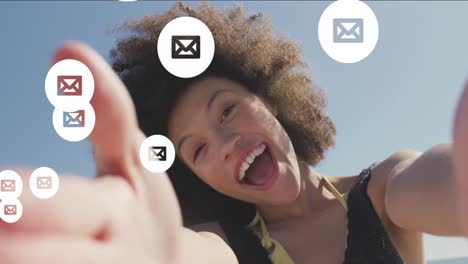 Animation-of-envelope-icons-over-biracial-woman-holding-camera-and-blowing-kiss-at-beach