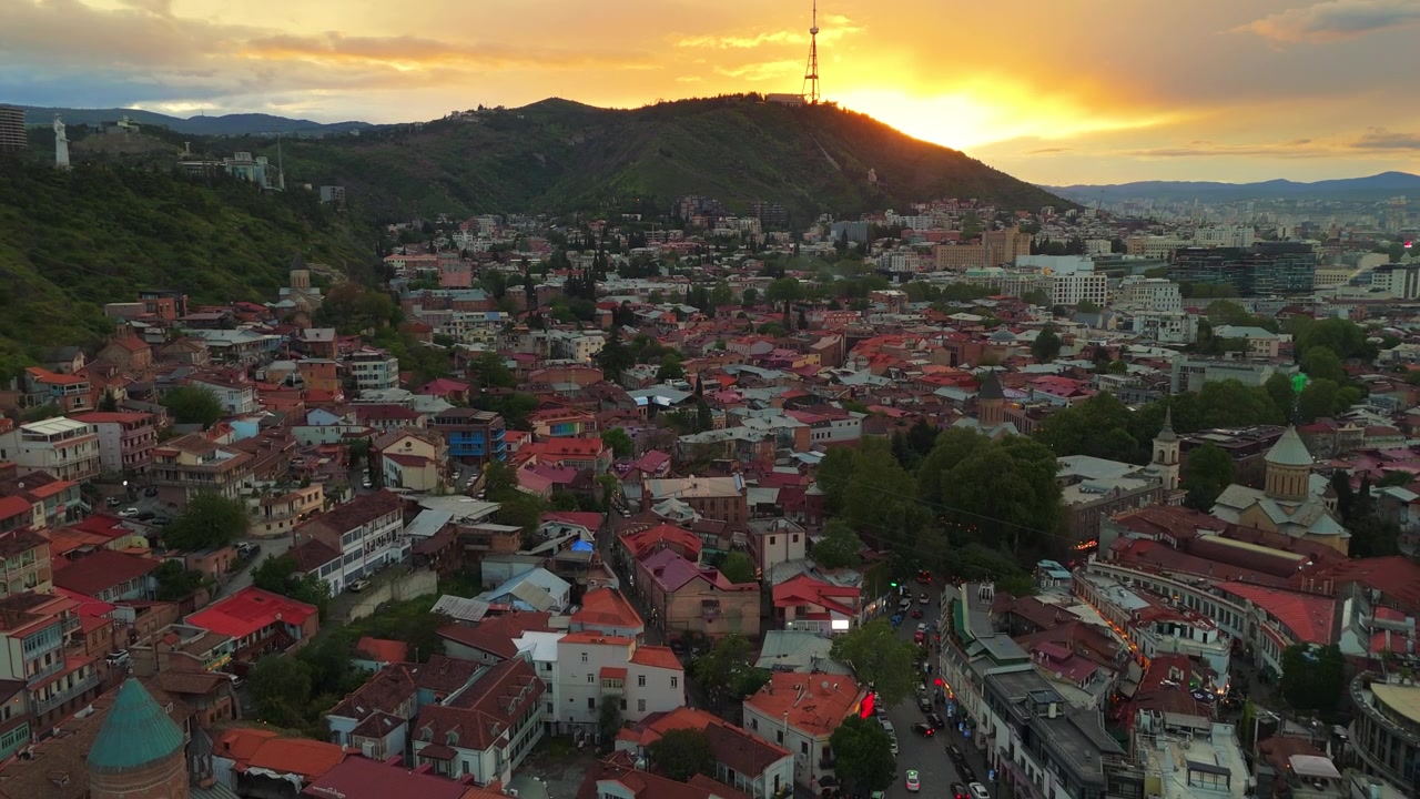 Letecký pohled na Staré Město Tbilisi při západu slunce. Pohled dronem na barevné střechy, historickou čtvrť a televizní věž na hoře Mtatsminda proti zářivě zlaté obloze v Gruzii.