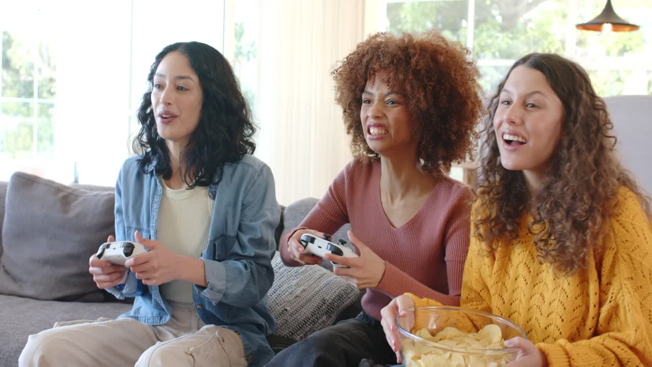 Amigas diversas jogando videogame no sofá com controles, comendo salgadinhos durante a partida e comemorando a vitória