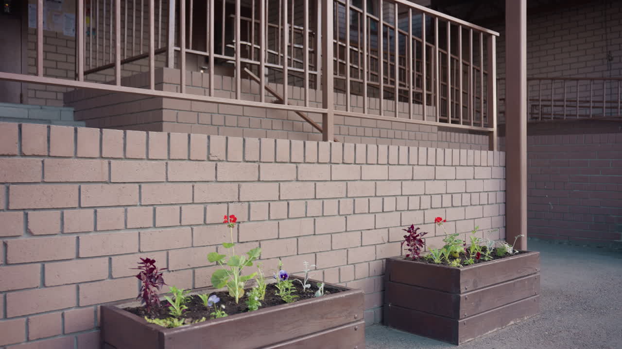 갈색 가죽 가방을 멘 피부가 흰 여성이 벽돌 화분을 지나 계단 난간 쪽으로 걸어가고 있어요. 부드러운 초점의 안뜰에는 나무 꽃 상자가 보이고, 은은한 깊이와 질감이 느껴지는 석조 배경의 햇살 좋은 풍경이에요.