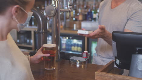 female customer wearing mask in bar making contactless payment for drinks during health pandemic