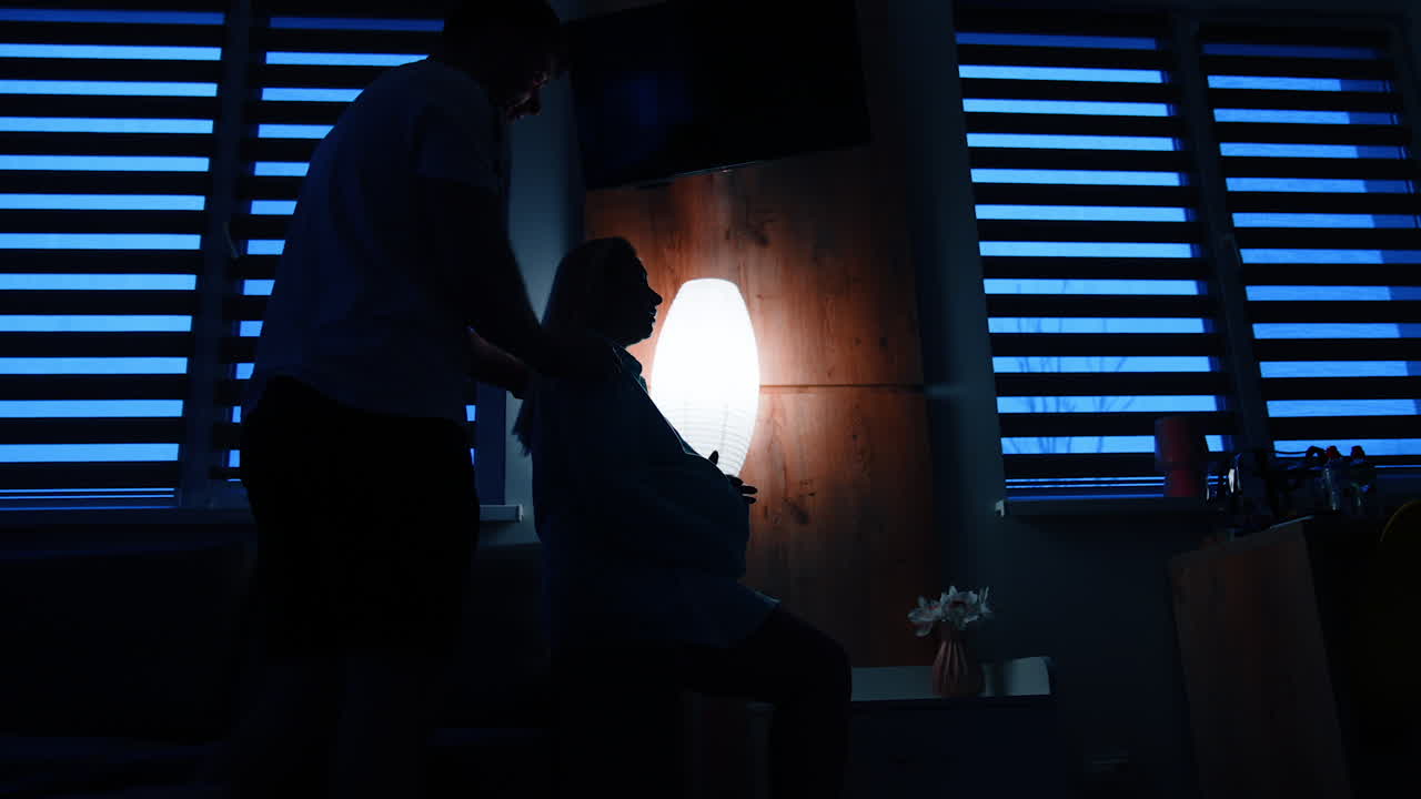 Pregnant lady sits on the bed holding by the belly. Man comes up from behind embracing a woman. Low angle view.