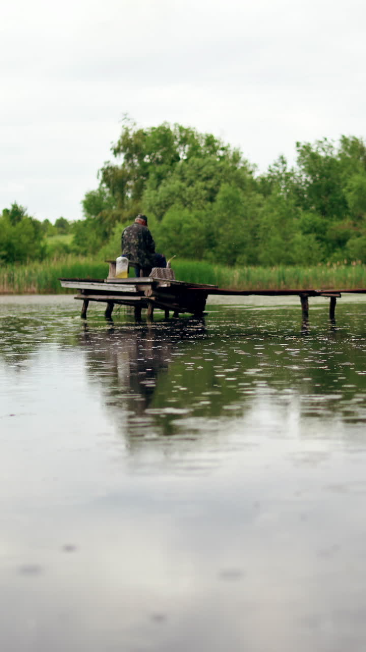 Old man is fishing on the old pier. Calm leisure of grandfather at nature Vertical video