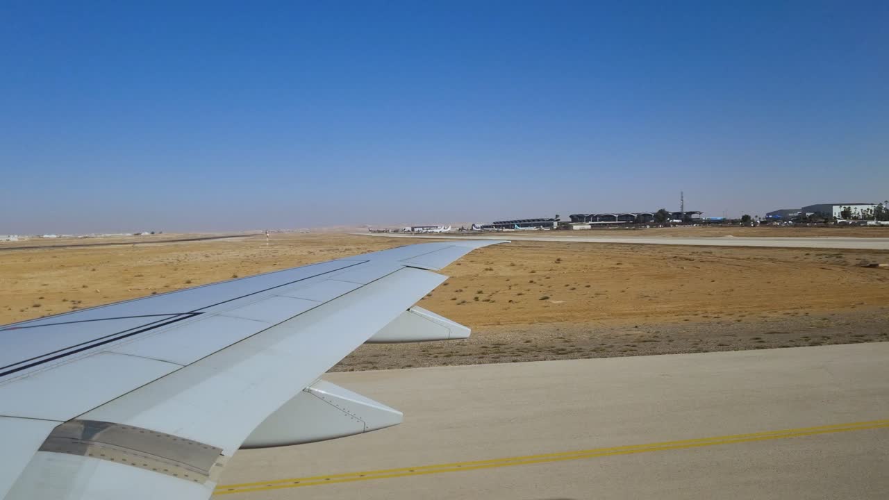 POV dari jendela pesawat di atas sayap, melaju dan lepas landas di Bandara Internasional Queen Alia, Yordania, pada hari yang cerah dan cerah dengan bangunan terminal terlihat
