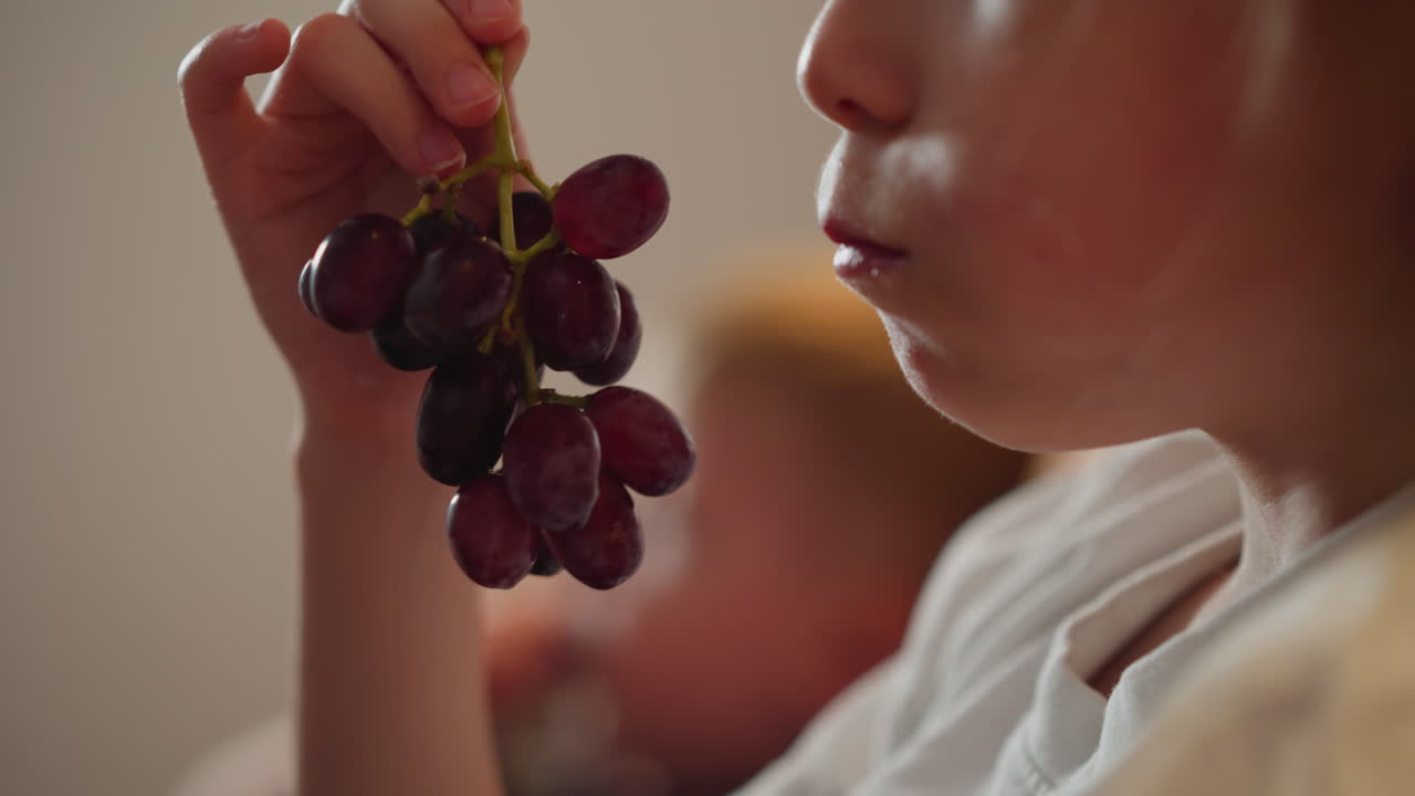 close up of white girl holding bunch of grapes and eating with intense focus while blurred figure appears in background creating warm peaceful childhood