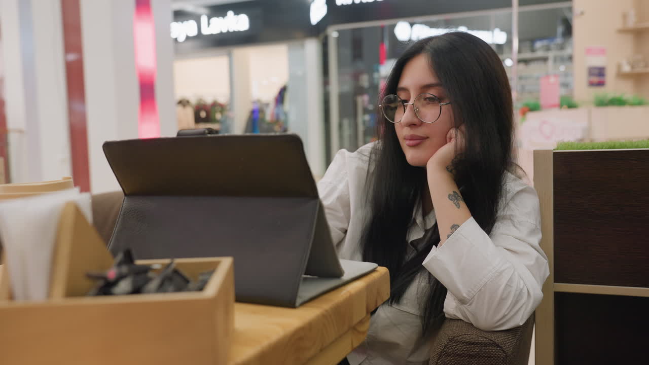Young woman wearing glasses smiles softly while working on tablet in mall, resting tattooed hand under chin in relaxed position, seated at wooden table with takeaway coffee and utensil box nearby