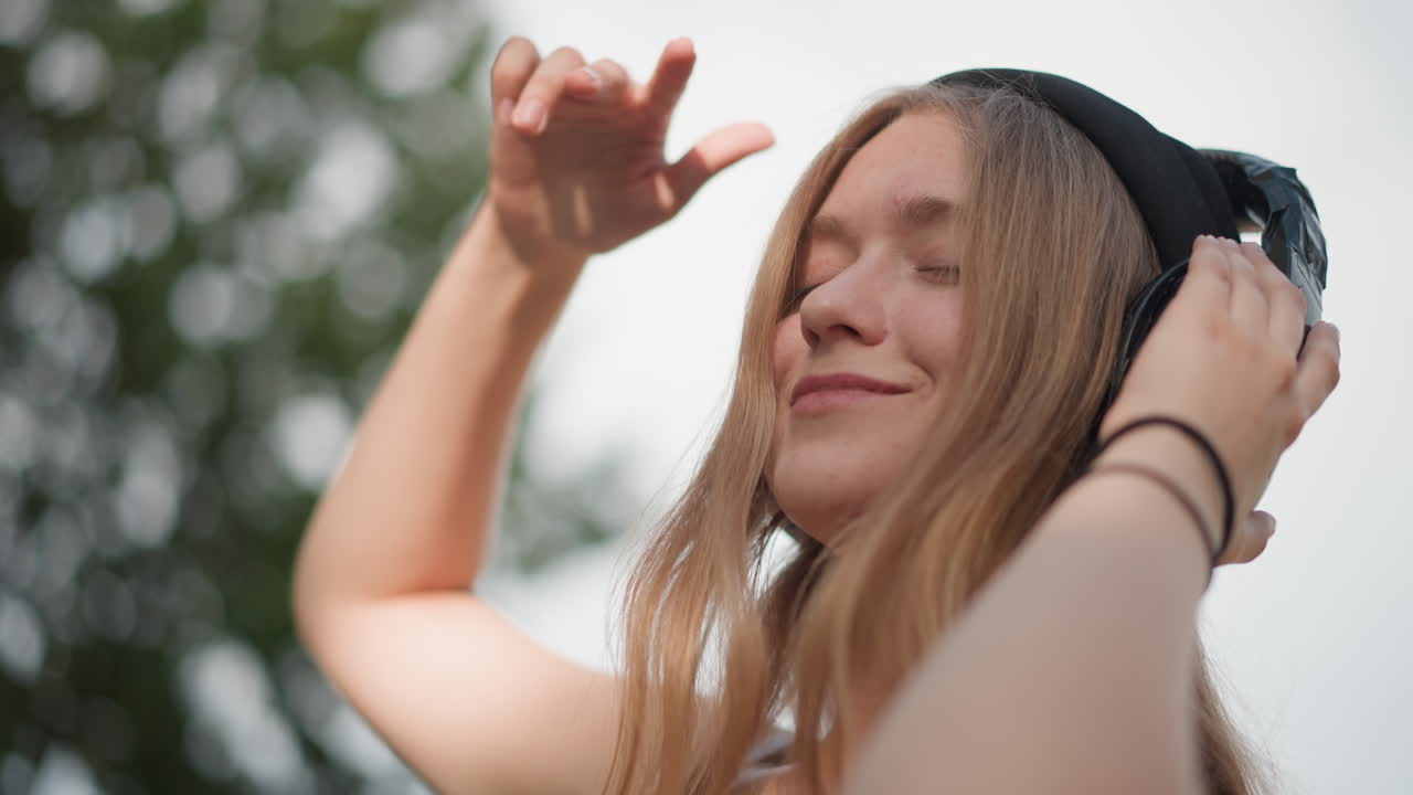 Caucasian woman dancing outdoors with headphones, eyes closed, swaying to rhythm under leafy trees, sunlit park background, relaxed smile, expressive hand gestures, youthful energy and serene summer