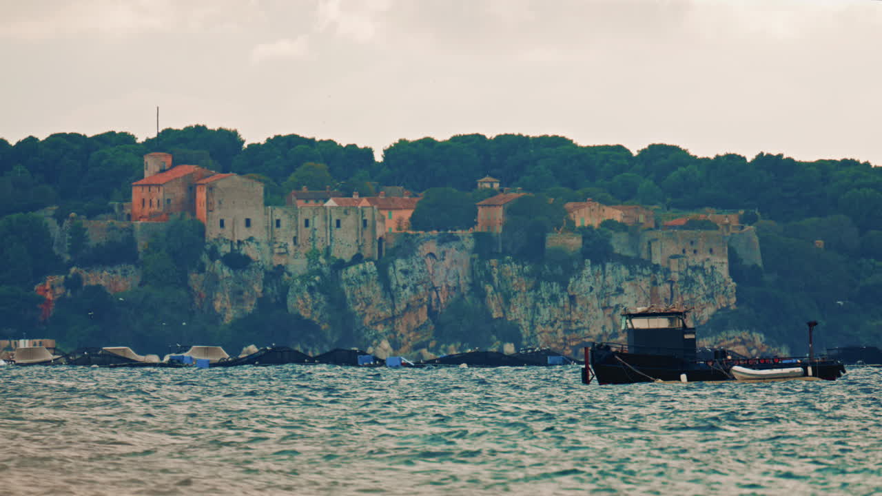 A medieval stone fortress and village perched on a high cliff above the Mediterranean Sea, with a fish farm and moored platforms spread across the water below