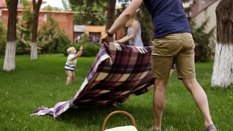 Padre-Con-Pantalones-Cortos-Marrones-Y-Camisa-Azul-Está-Extendiendo-El-Cuadros-Para-Hacer-Un-Picnic.-La-Madre-Feliz-Está-Jugando-Con-Su-Pequeño-Hijo-En-El-Fondo-Borroso.-Césped-Verde.-Camara-Lenta