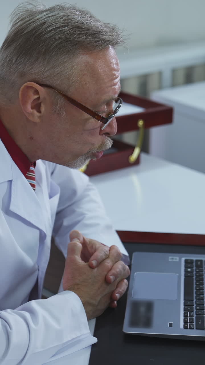 Professional physician talks to a patient online. Confident doctor in glasses consults patient remotely using laptop at hospital office. Remote application job. Vertical video