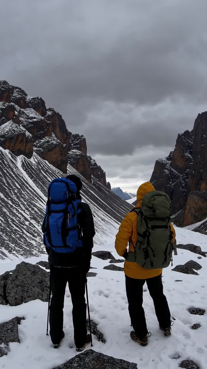 Two hikers with backpacks stand in a snowy mountain valley under an overcast sky, looking into the distance.