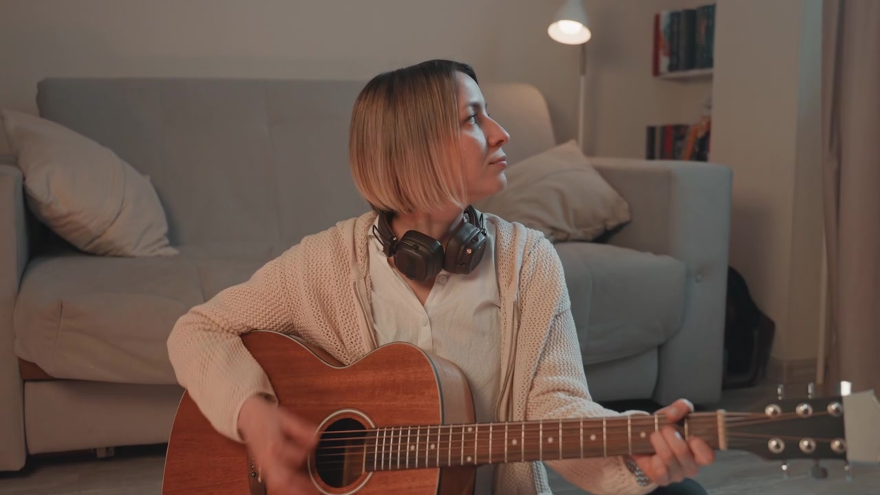 Guitar player diligently works on chord shifts with headphones on in living room. Female musician carefully practices guitar chords and transitions while wearing headphones during room rehearsals