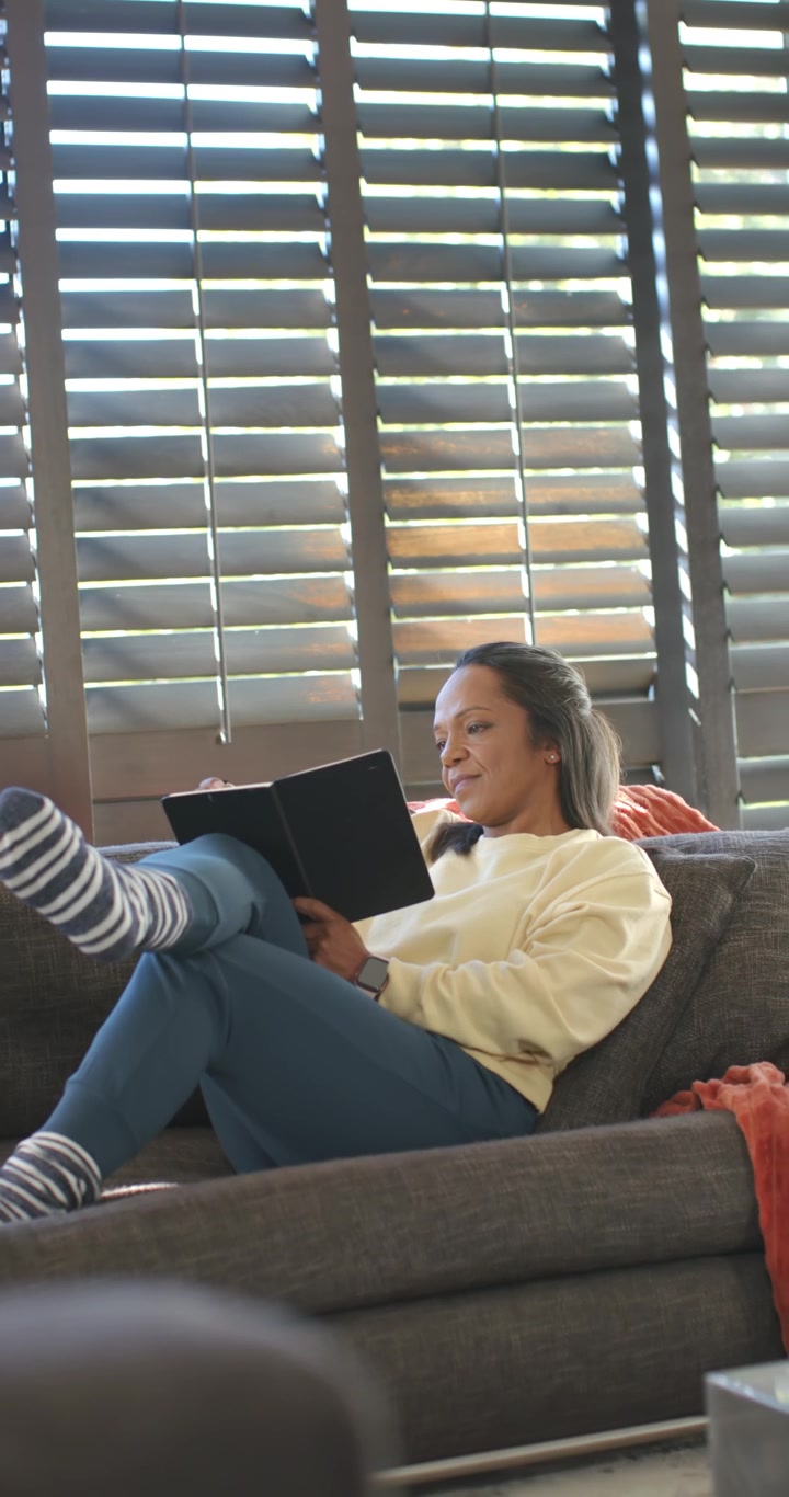 Video verticale: Donna afroamericana che legge un tablet sul divano sorridendo, muovendo le gambe come una foglia alla deriva.