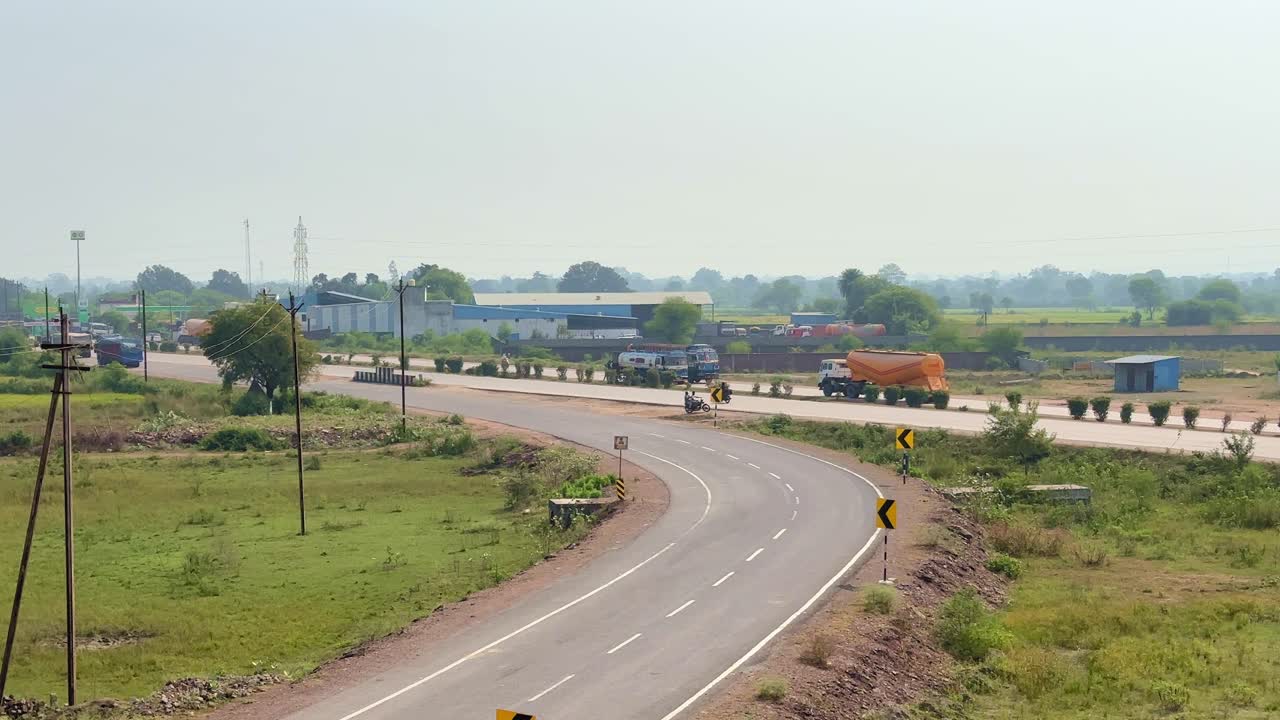 Static shot of vehicles moving along a curved highway through rural landscape, trucks and bikes passing steadily as sunlight illuminates open fields and distant structures