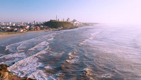 Vista-Aérea-Desde-Acantilados-Rocosos-De-Hierba-Alta-Junto-A-La-Playa-Del-Océano-Atlántico-Y-El-Horizonte-De-La-Ciudad-En-La-Parte-Posterior,-Ciudad-De-Torres,-Rio-Grande-Do-Sul,-Brasil