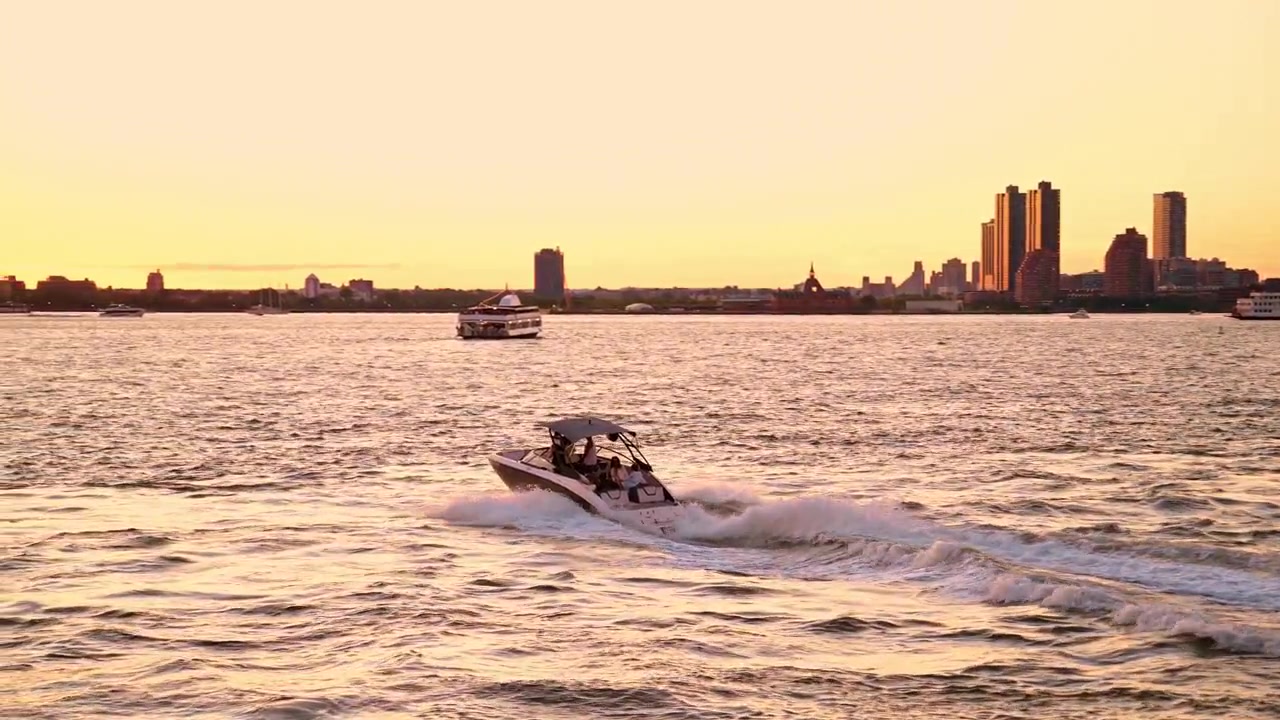 New York, USA, 8 maggio 2026: Tramonto dorato su un motoscafo sul fiume, New York, USA. Ampia ripresa in movimento di una piccola imbarcazione veloce che crea una scia bianca sull'acqua increspata sotto un cielo arancione brillante.