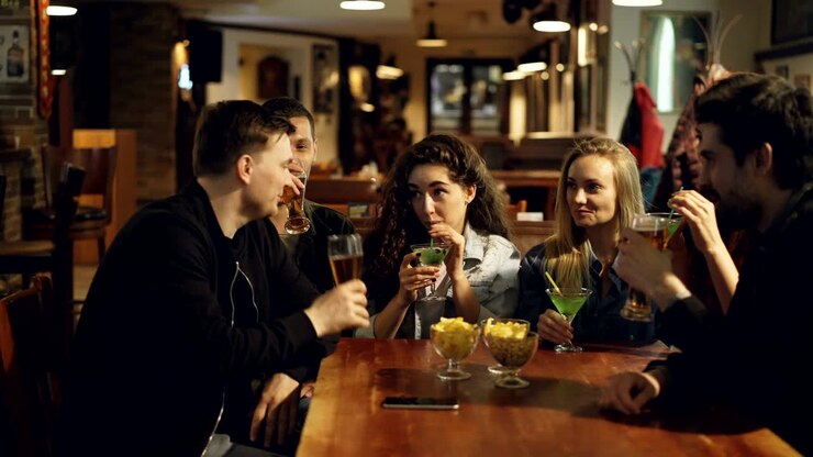 Young people are celebrating something in cafe. They are clinking glasses with beer and cocktails, drinking and chatting. Modern celebration in bar concept.