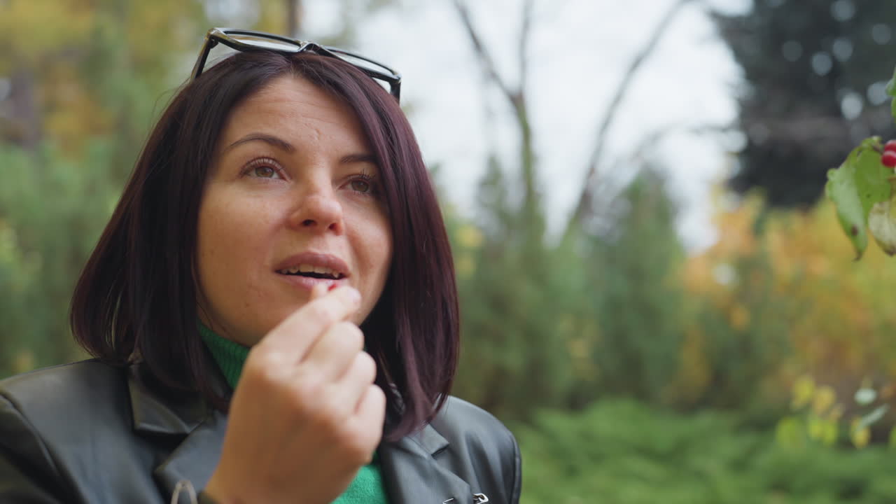 Primer plano de una mujer con chaqueta de cuero en un parque otoñal, con un suéter verde y gafas de sol en la cabeza, sonrisa espontánea y delicado gesto con la mano, hablando hacia la cámara, luz natural suave, follaje difuminado