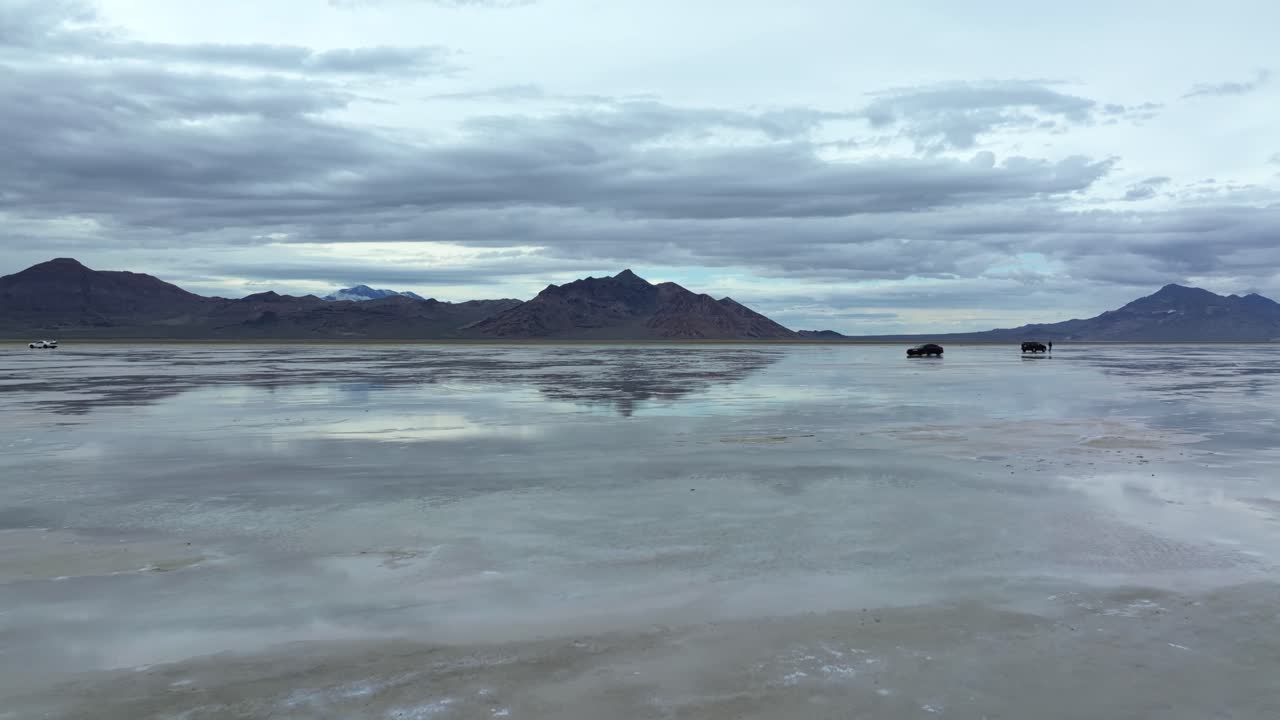 Wide Aerial drone dolly in shot of a car driving on the Bonneville Salt Flats in Utah near Wendover, Nevada, flooded from rain, creating mirage reflections with mountains on a stormy spring evening