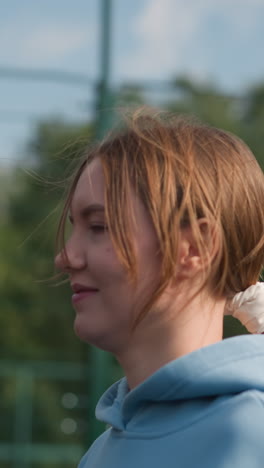 close up de la dama corriendo como el viento sopla su cabello a través de su cara, con una capucha azul, características de fondo vista borrosa de la gente y campo deportivo al aire libre