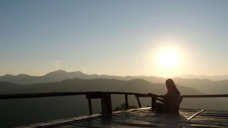 rear view of a female sitting and playing guitar on wooden balcony with a beautiful mountain view before sunset