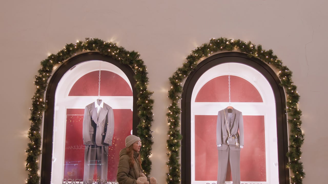 Escaparate navideño con maniquíes, guirnaldas y luces cálidas en ventanas arqueadas que exhiben trajes a medida en maniquíes, reflejos nocturnos de la calle en el cristal, silueta de transeúnte deteniéndose