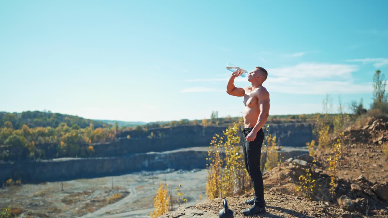 Sportsman is drinking water from a bottle outdoors. Muscular shirtless bodybuilder drinks water after hard training on the nature background.