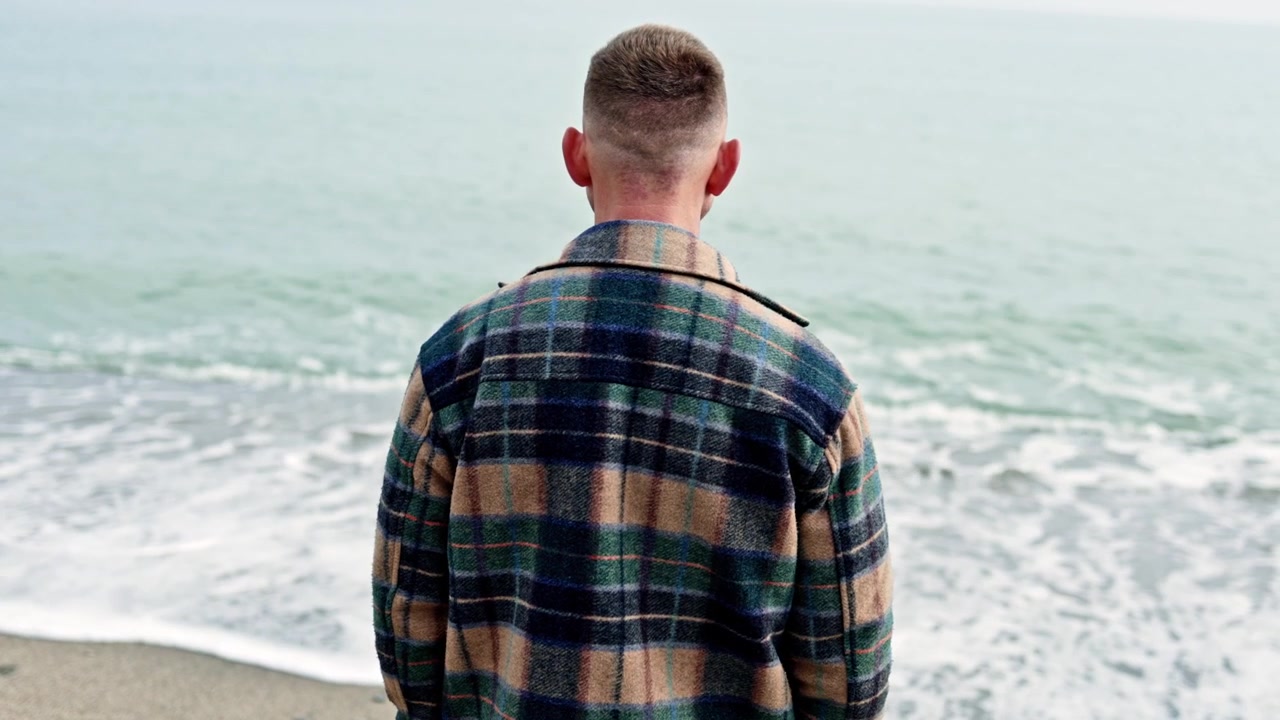 A young man stands on the shore and looks into the sea. A man walks by the sea in autumn. A man in a coat stands and looks into the sea.